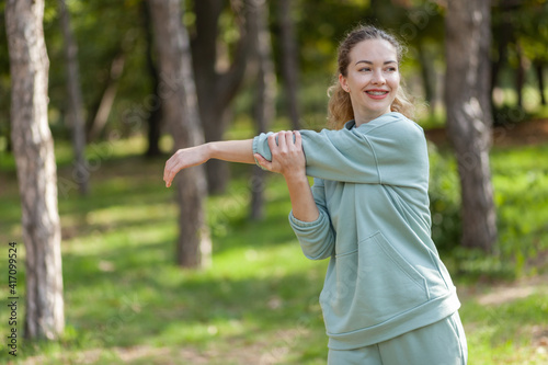 Attractive fit slim Caucasian woman with curly hair in sportswear is doing stretching exercise in the park or forest on a bright sunny day. Healthy lifestyle