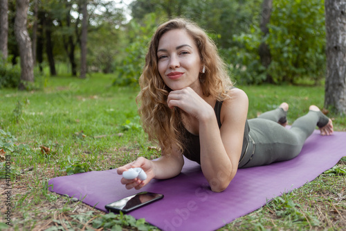 Attractive young fit woman with curly hair is holding wireless headphones and getting ready to listen to music while lying on the mat. © splitov27