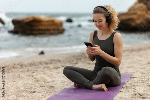 Attractive caucasian woman yogi listening to music with headphones and uses smartphone, relaxing while sitting on mat at the beach