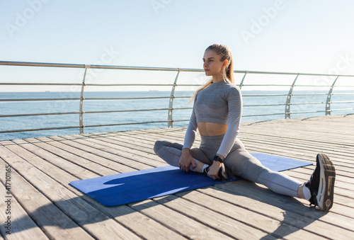 Fit woman in sportswear doing stretching while sitting on the mat on the beach at bright sunny day. Healthy lifestyle