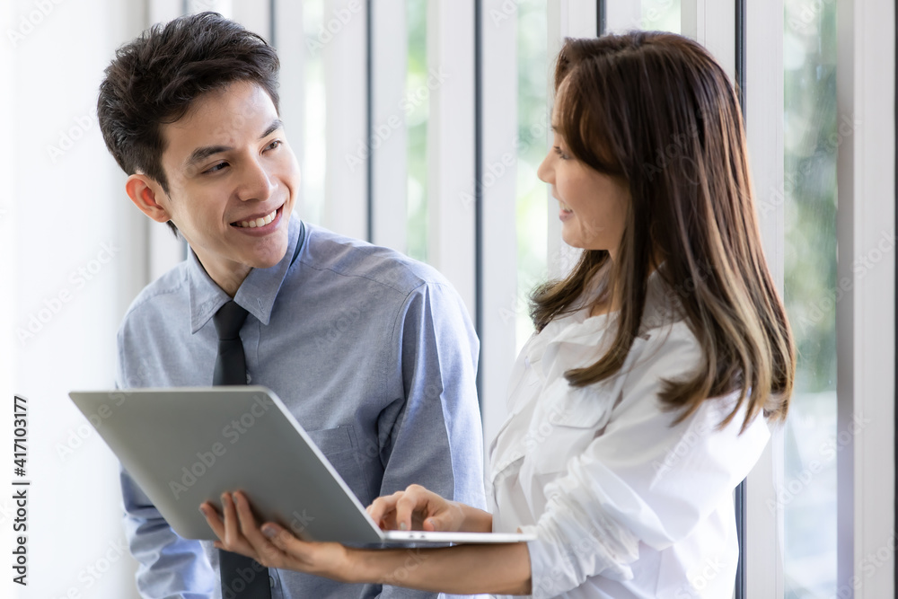 Obraz premium Beautiful black long hair Asian businesswoman standing watching the screen of laptop notebook computer and talking with businessman with intimate manner in office