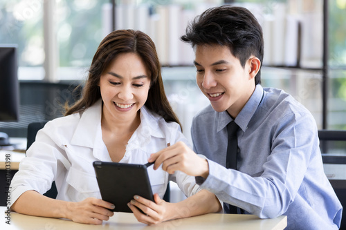 Beautiful black long hair Asian businesswoman and gook looking cute Asian businessman sitting together and watching the screen of tablet computer in office with smile face