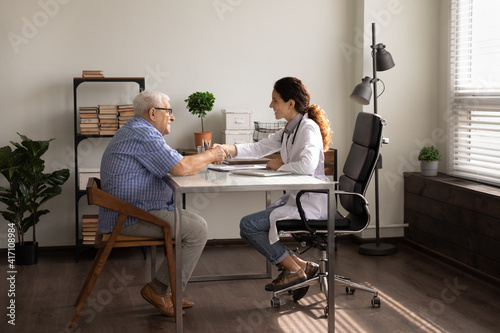 Side view smiling female therapist and mature patient shaking hands, greeting, sitting at table in doctor office, celebrating good medical checkup results, client signing health insurance contract © fizkes