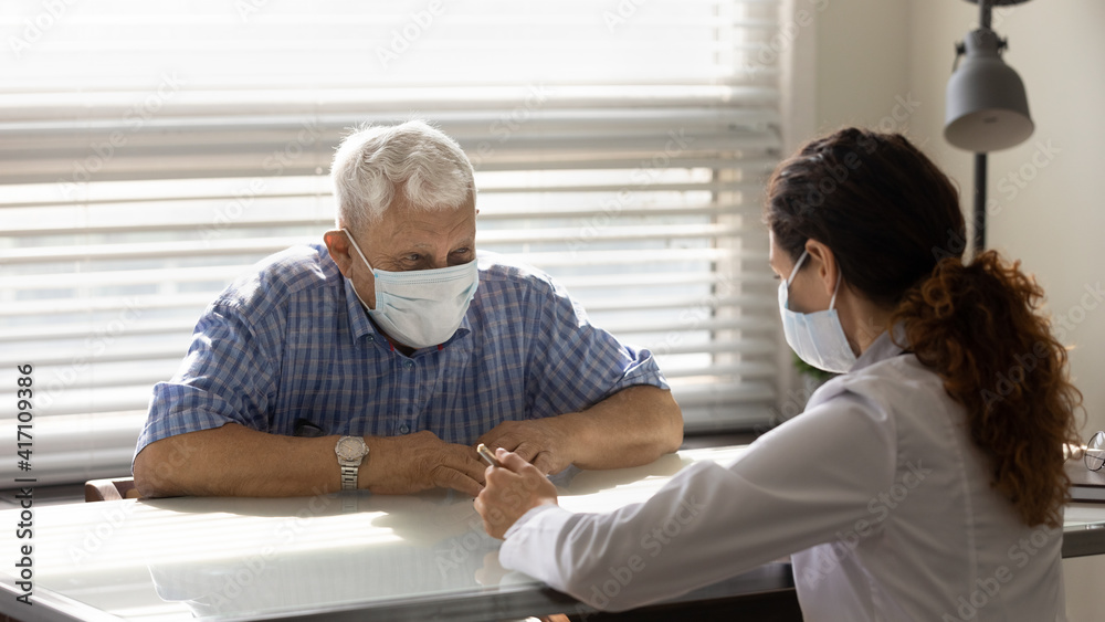 Obraz premium Close up female doctor consulting smiling older patient wearing medical face mask about health insurance agreement at meeting in hospital, therapist and elderly patient discussing contract terms