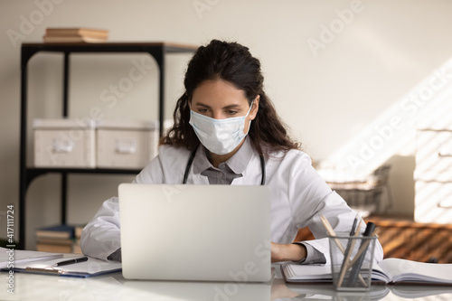 Close up confident professional female doctor wearing protective face mask and uniform using laptop, focused physician looking at screen, working in hospital office, consulting patient online