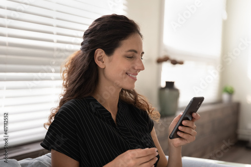 Close up smiling woman looking at phone screen, having fun with device, chatting with boyfriend or shopping online, young female holding smartphone, sitting on couch at home, enjoying leisure time
