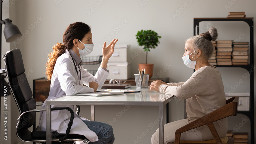 Side view female doctor wearing face mask and uniform consulting mature patient about treatment at meeting in hospital office, therapist and senior woman discussing checkup results, healthcare Side view female doctor wearing face mask and uniform consulting mature patient about treatment at meeting in hospital office, therapist and senior woman discussing checkup results, healthcare