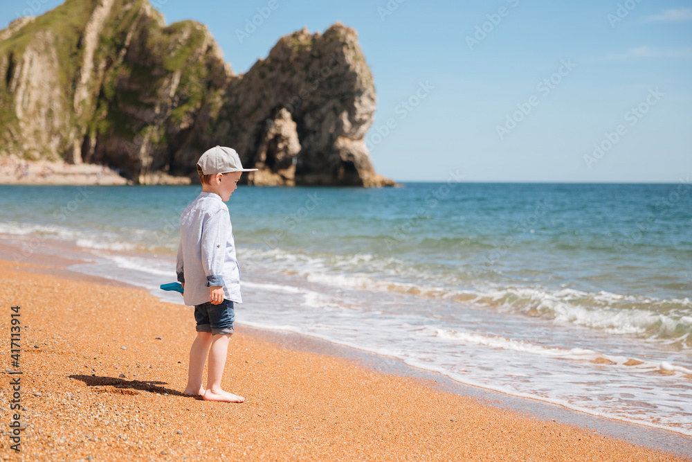 Little boy with capy and a shovel plays on a pebble beach. Child on a beautiful beach. Beach in England. Summer vacation in Germany 