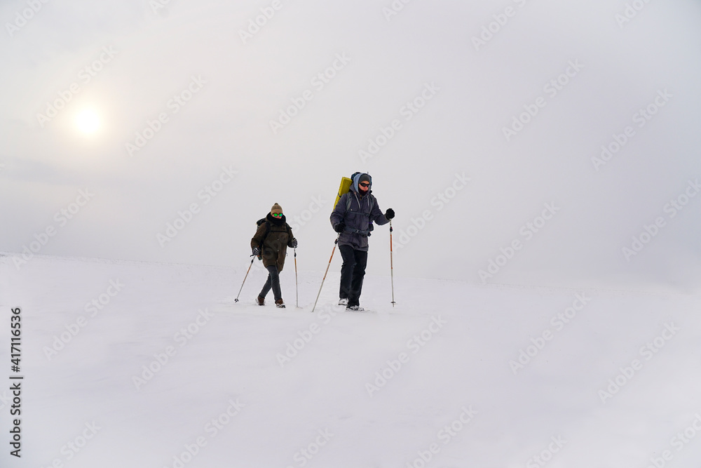 Two guys walk through loose snow during a winter expedition. They carry large backpacks, warm jackets. They hold trekking sticks in their hands.
