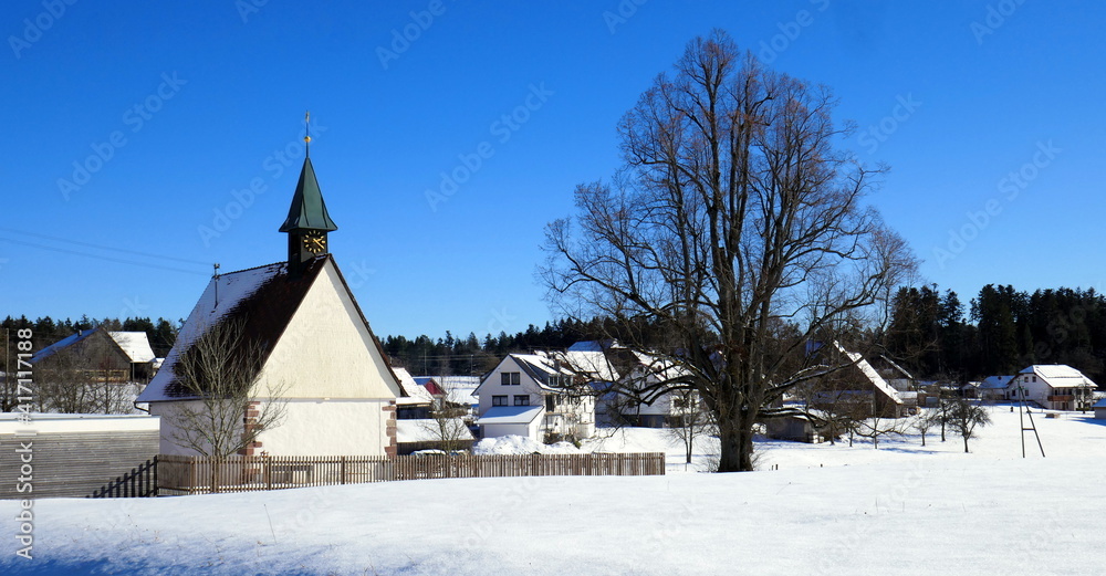 kleines Schwarzwalddorf im Winter mit Kirche, Häusern und kahlem Baum unter blauem Himmel