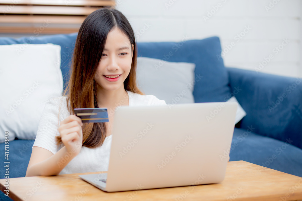 Young asian woman sit on sofa using laptop computer shopping online with credit card buying to internet, happy girl payment with e-business on couch, purchase and payment, business concept.