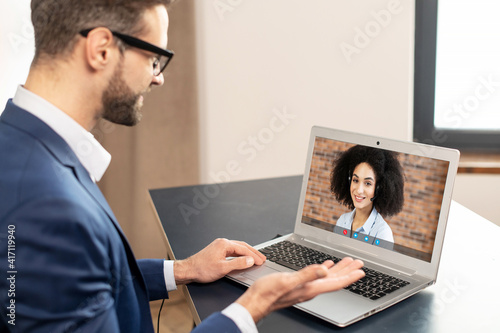 An intelligent young male office worker in formal wear and glasses sitting at the desk at home office, having a virtual online meeting on laptop and talking listening to his female colleague or friend