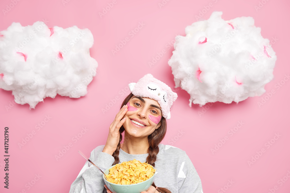 Pretty gentle woman tilts head smiles toothily touches her soft healthy skin after beauty procedures wears pink collagen patches under eyes poses with bowl of cornflakes has breakfast in morning Pretty gentle woman tilts head smiles toothily touches her soft healthy skin after beauty procedures wears pink collagen patches under eyes poses with bowl of cornflakes has breakfast in morning