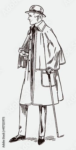 Standing man from the early 20th century wearing inverness cape overcoat and hat, holding walking stick