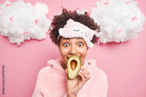 Positive surprised dark skinned young woman covers mouth with hald of avocado going to have beauty procedures and apply mask after awakening dressed in soft comfortable slumber suit poses indoor.