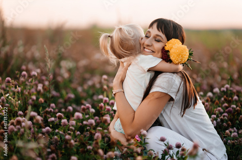 Happy family, mother and child daughter are hugging on a flower meadow in summer nature at sunset time