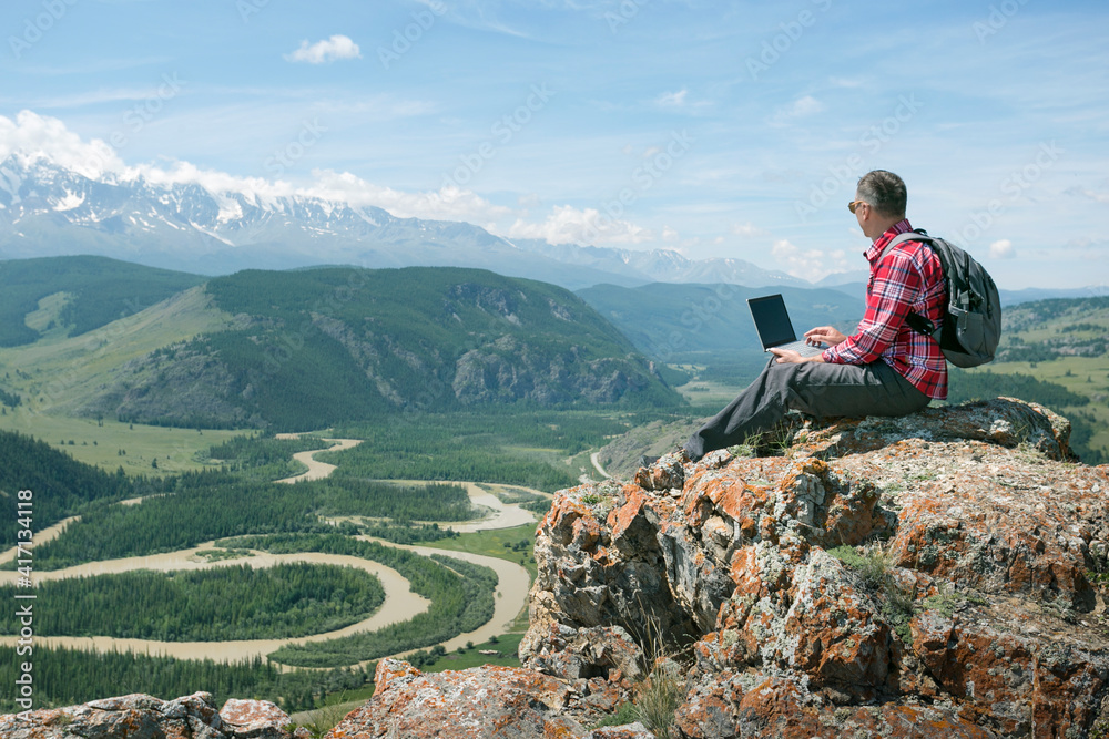 Adult man working outdoors with laptop sitting in mountains. Concept of remote work or freelancer lifestyle.
