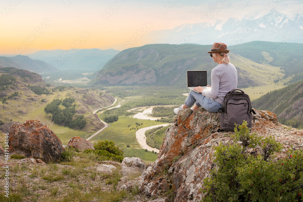 Fototapeta premium Adult woman working on her computer on the top of the mountain at sunset. Remote work concept.