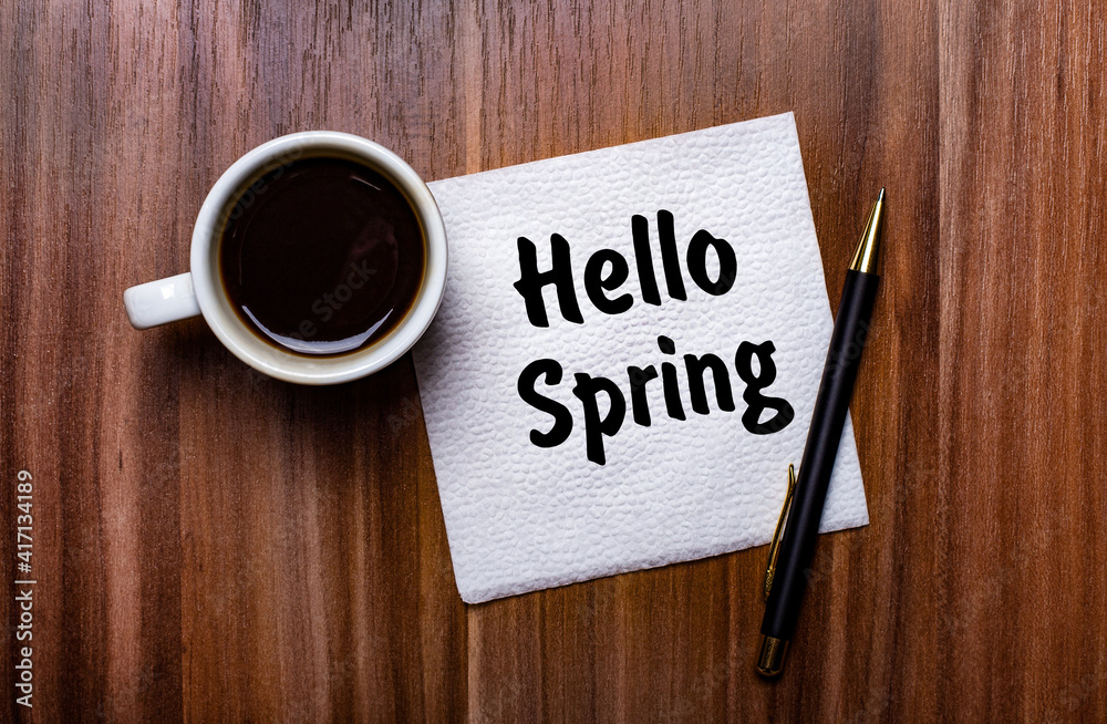 On a wooden table next to a white cup of coffee and a pen is a white paper napkin with the words HELLO SPRING On a wooden table next to a white cup of coffee and a pen is a white paper napkin with the words HELLO SPRING