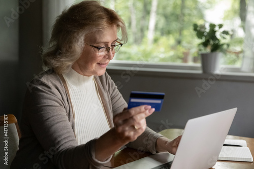 Happy mid adult 50s aged customer using credit card and laptop. Female mature retiree making payments or transactions, booking online. Pensioner shopping and buying goods on internet stores