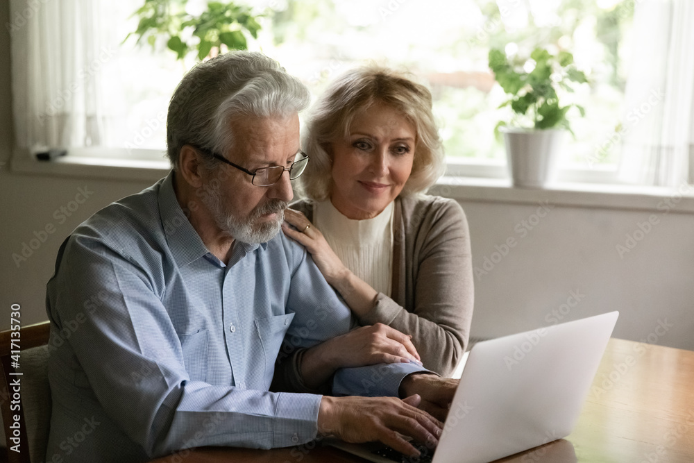 Happy confident mature family couple using app on computer at home. Senior husband and wife typing on laptop, checking bank account or finance insurance balance, shopping, reading or booking online
