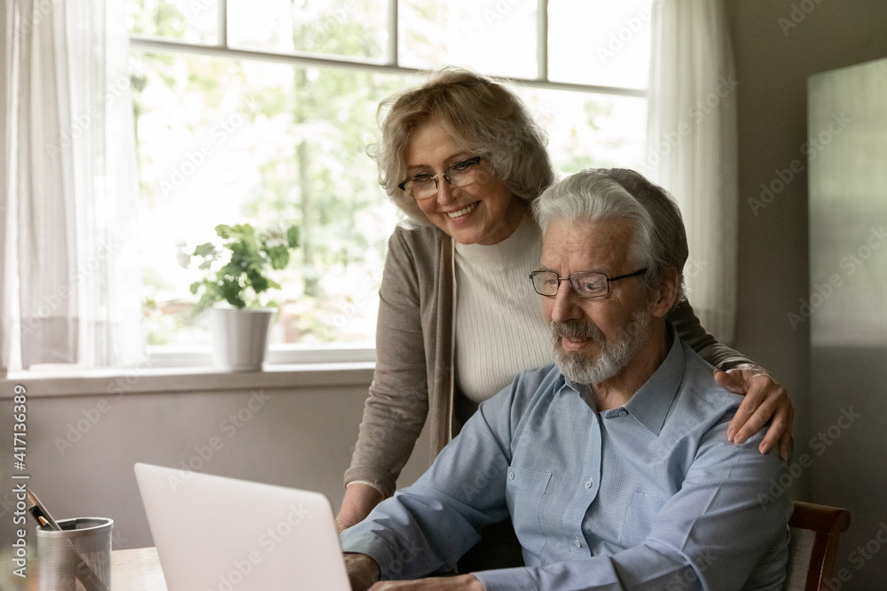 Happy mature OAP couple study internet app on laptop, grandparents use computer at home for online shopping. Smiling middle aged pensioner wife hugs senior husband during video call to relations