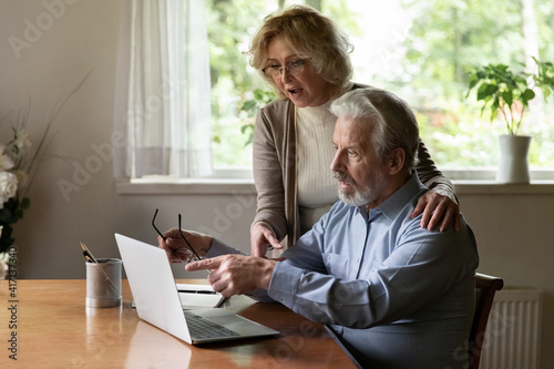 Senior couple using app at home, studying new online app on modern tech device together. Mature man pointing at laptop screen, reading unexpected news, showing content to surprised middle aged wife © fizkes