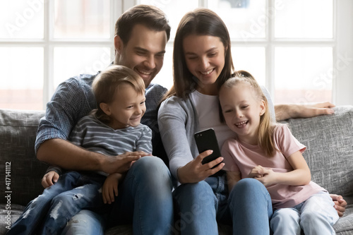 Happy parents and z generation kids make video phone call from home. Family rests on living room sofa. Couple hugging children, using internet app on smartphone, playing online funny game, having fun