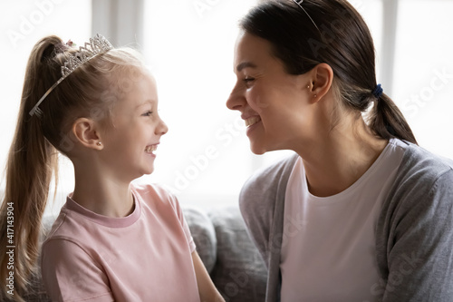 Happy mom expressing love to little daughter girl wearing princess diadem. Mother talking to child, smiling, laughing, having fun with cute little kid at home. Family bonding, relationship concept