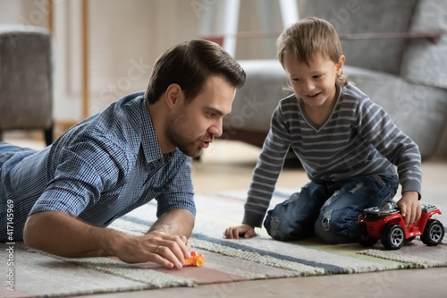 Happy dad and son wheeling toy cars on heat floor in living room. Father and gen Z boy lying on carpet, playing together at home, enjoying activity, games, leisure time, family active playtime