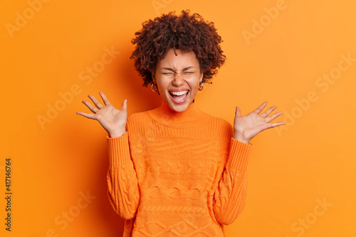 Positive overjoyed young African American woman raises palms feels very glad expresses joy dressed in casual jumper isolated over orange background. People emotions happiness concept. Monochrome photo