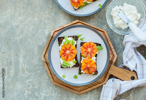 A healthy breakfast or snack, open sandwiches on rye bread with cottage cheese or cream cheese, slices of bloody oranges and herbs on two plates on a gray concrete background.