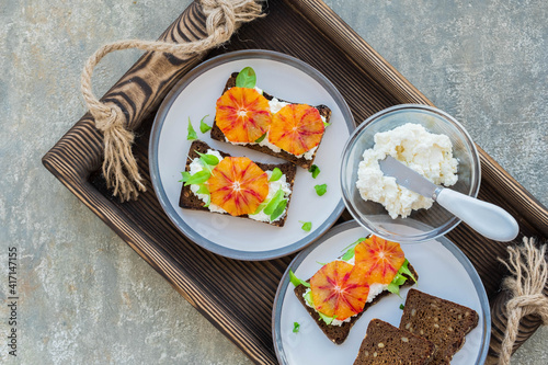 A healthy breakfast or snack, open sandwiches on rye bread with cottage cheese or cream cheese, slices of bloody oranges and herbs on two plates on a gray concrete background.
