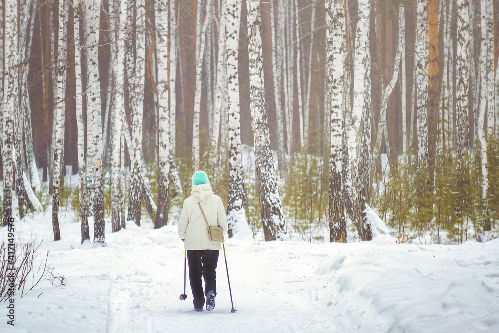 A woman aged in the winter forest is engaged in Scandinavian walking.  active lifestyle in old age.  health promotion.  woman in winter forest.  jogging in the fresh air among the birches.