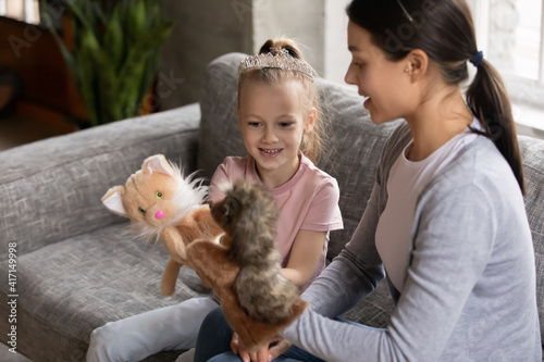 Happy mum and excited daughter girl wearing princess tiara talk play toy fluffy hand puppets together at home. Mother or nanny entertains preschool kid with role game activity. Family, daycare concept photo