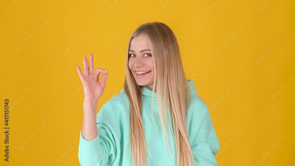 Cute young blonde girl looking at camera charming smile showing ok okay gesture isolated on yellow background. People lifestyle concept. Cute young blonde girl looking at camera charming smile showing ok okay gesture isolated on yellow background. People lifestyle concept.