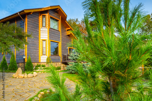 Coniferous tree in front of cottage. Two-storey house on a summer day. Landscape of territory near cottage. Decoration of territory next to house. Landscape design services. Designer services.