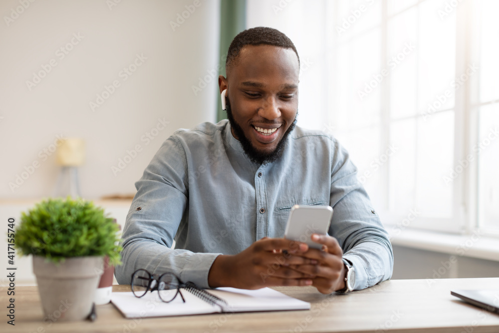 Black Businessman Using Smartphone And Earbuds At Workplace In Office Black Businessman Using Smartphone And Earbuds At Workplace In Office