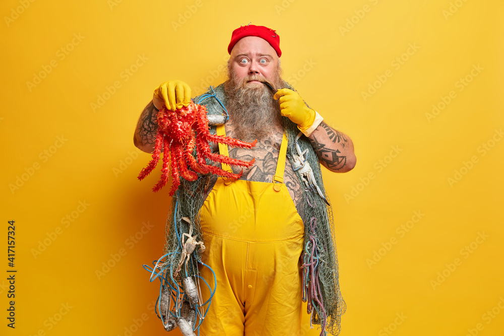 Experienced sailor with thick beard smokes pipe didnt expect to catch so big octopus wears hat rubber gloves and overalls has trip around world isolated over yellow background. Wanderlust concept