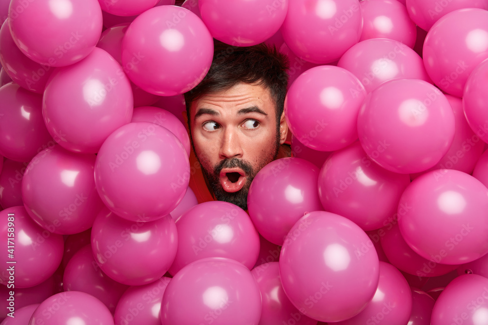 Obraz premium Surprised bearded European man sticks head through decorated background with inflated balloons prepares for holiday being shocked by unexpected relevation. People celebration emotions concept