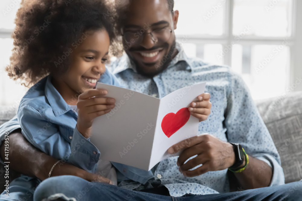 Happy little daughter showing handmade postcard to smiling dad, reading wishes. Family celebrating fathers day. Girl giving surprise greeting card with hand drawn love heart to daddy. Close up