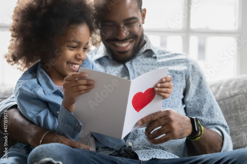 Happy little daughter showing handmade postcard to smiling dad  reading wishes. Family celebrating fathers day. Girl giving surprise greeting card with hand drawn love heart to daddy. Close up
