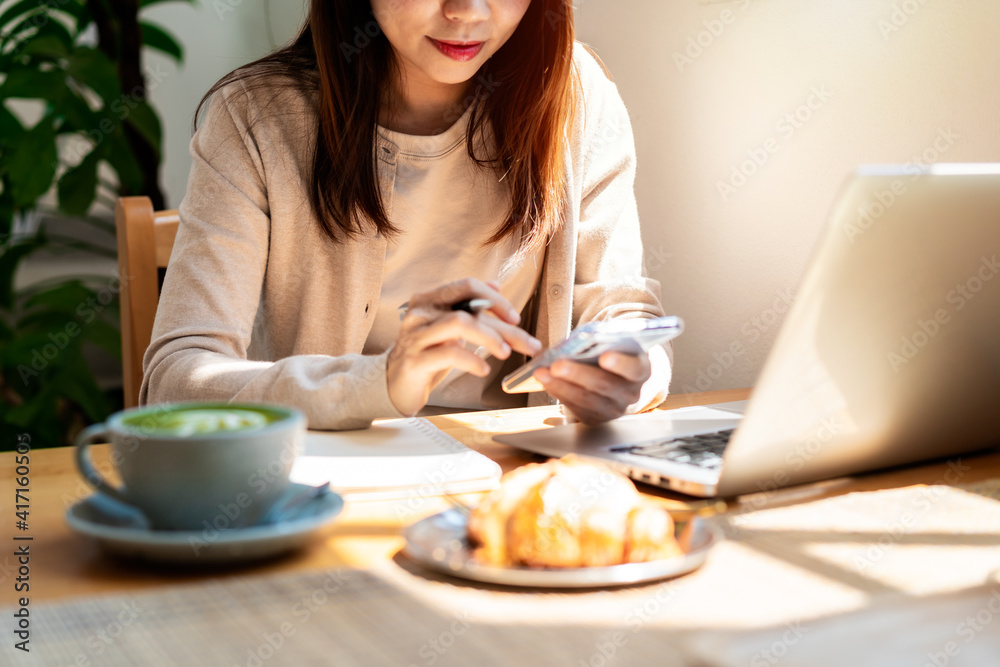 Young woman with cup of coffee using mobile phone and working on laptop at coffee shop