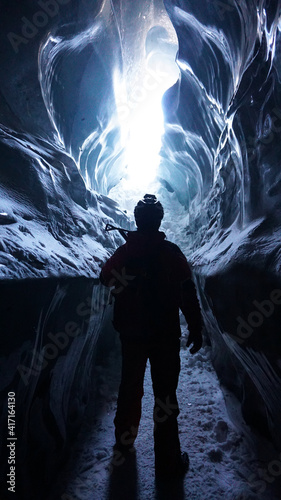A climber with an ice pick stands at the exit of an ice cave. Inside Bogdanovich Glacier. High ice walls are sometimes covered with snow and stones lie. A flashlight is shining. Mountains of Almaty