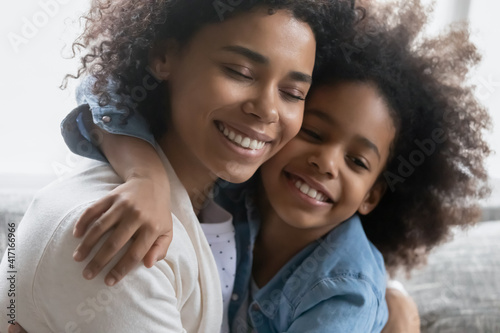 Happy African American girl embracing her peaceful mum with closed eyes. Mother enjoying leisure time with her daughter kid at home, hugging child and cuddling child. Family bonding, love concept