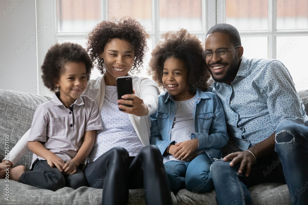 Happy black family couple and two kids taking selfie picture on smartphone at home. Millennial African American parents and children using cell phone for video call and virtual chat with grandparents