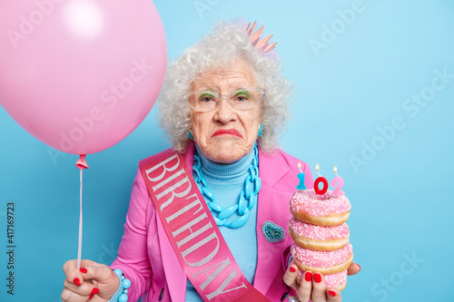 Portrait of curly displeased wrinkled woman frowns face has bright makeup dressed in festive clothes celebrates 102nd birthday holds sweet tasty doughnuts inflated balloon isolated over blue wall