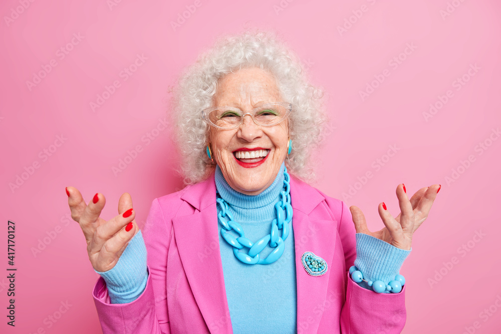 Happy senior curly woman enjoys life in old age raises hands smiles positively dressed in fashionable outfit wears bright makeup has red manicure wrinkled face isolated over pink background. Happy senior curly woman enjoys life in old age raises hands smiles positively dressed in fashionable outfit wears bright makeup has red manicure wrinkled face isolated over pink background.