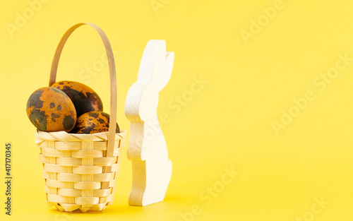 Easter colored quail eggs in a basket and a wooden rabbit on a yellow background. Easter holiday concept. Copy space, close up