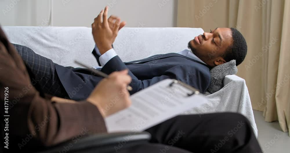 Afro american business man lies on couch in office of psychologist consults with psychiatrist going rehabilitation psychotherapy speaks with specialist unrecognizable woman writing notes in notebook Afro american business man lies on couch in office of psychologist consults with psychiatrist going rehabilitation psychotherapy speaks with specialist unrecognizable woman writing notes in notebook